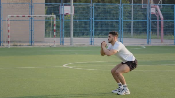 Młody energiczny Hiszpan Arab w letniej odzieży sportowej pokazuje technikę squat. Bliskowschodni indyjski sportowiec, piłkarz lub biegacz rozgrzewa się podczas treningu na stadionie, robi ćwiczenia cardio — Wideo stockowe