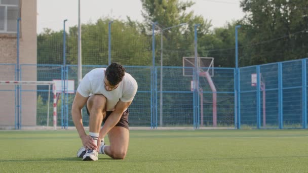 Middle Eastern Indian sportsman runner or footballer in summer sportswear feels soreness, discomfort during workout at stadium. Suffering guy holding on ankle, rubs bruised area, got sports leg injury — Stock Video