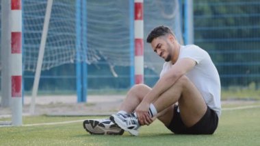 Acılar içindeki Orta Doğulu futbolcu kaleye karşı çimenlerde oturuyor, bileğini tutuyor, milenyum atleti yaz sporu giysisinde yaralanmış, yaralanmış, devam edemiyor.