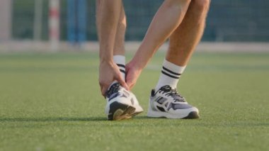 Beyaz spor ayakkabılarıyla güçlü erkek bacaklarına ve spor stadyumunun çimlerinde çoraplarına yakın çekim. Acı çeken bir futbolcu ayak bileğini tutuyor, yaz sporu kıyafetleri giyen bir atlet ezilmiş bölgeyi ovuyor, spor bacağından sakatlanıyor.