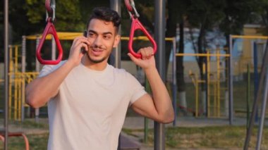 Spor giyimli Hintli erkek öğrenci sabah antrenmanı sırasında telefona bakar. Stadyumdaki spor malzemelerini kullanarak gülümseyen Orta Doğu Arap milenyum sporcusu hücresel konuşmalar yapıyor.