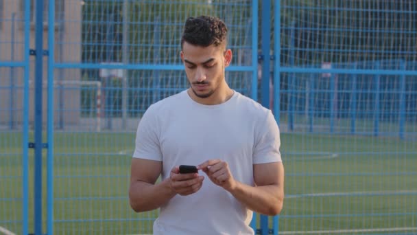 Сосредоточенный спортсмен индийский парень в белой футболке, спортивная одежда смотрит на секундомер, устанавливает приложение для отслеживания электронной активности. Ближневосточный спортсмен использует функции смартфона для мониторинга результатов тренировок — стоковое видео