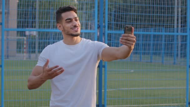 Jovem blogueiro esportivo indiano em sportswear olhando para câmeras de smartphones grava transmissões do estádio, se comunica com o público. Atleta do Oriente Médio usa celular para videoconferência — Vídeo de Stock
