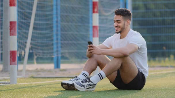 Glimlachend Arabisch voetballer uit het Midden-Oosten zittend op gras van voetbalveld tegen doel, met smartphone kijkend naar het leesbericht op het mobiele scherm. Millennial Indiase atleet kreeg goed nieuws voelt zich gelukkig — Stockvideo