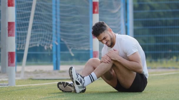 Nieszczęśliwy ranny bliskowschodni piłkarz siedzący na trawie boiska do piłki nożnej przeciwko bramce, trzymający kolano. Tysiąclecia sportowiec w odzieży sportowej cierpi na ból nóg doświadcza kontuzji, nie może kontynuować — Wideo stockowe