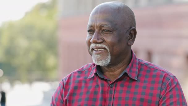 Elderly black man looks pensively aside, slowly turns head looking at camera. Portrait handsome pensioner male face closeup. Friendly mature bearded african american smiling grandfather in shirt — Stock Video