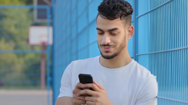Joven atleta árabe de Oriente Medio en ropa deportiva de verano sosteniendo el teléfono móvil, utilizando la aplicación virtual, servicio en línea en el teléfono inteligente, mensajes de texto, mensaje de escritura de chat, pantalla táctil, de pie en el estadio — Vídeo de stock