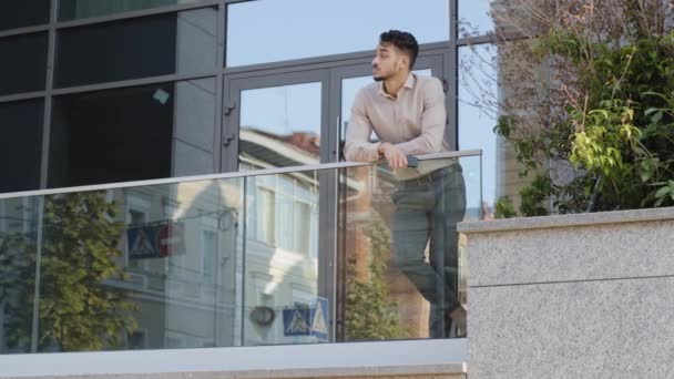 Joven hombre de negocios hispano confiado parado en barandilla edificio de oficinas rascacielos moderno mirando hacia otro lado sosteniendo el teléfono pensando descansando. Exitoso líder hombre en balcón terraza relajante pensar — Vídeos de Stock