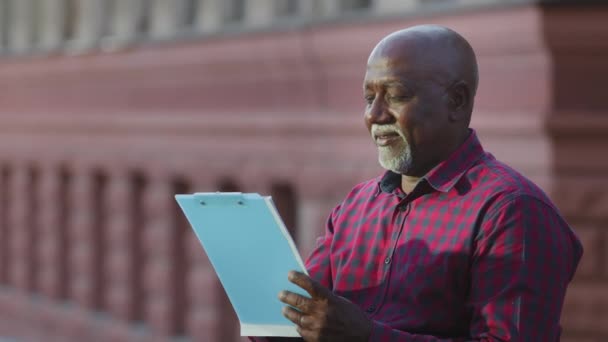 Érett felnőtt afro-amerikai férfi szakember áll a szabadban papírvágólappal. Utazó, építész vagy művész épületeket vizsgál, építészetet elemez, rajzokat készít a nyári séta alatt — Stock videók