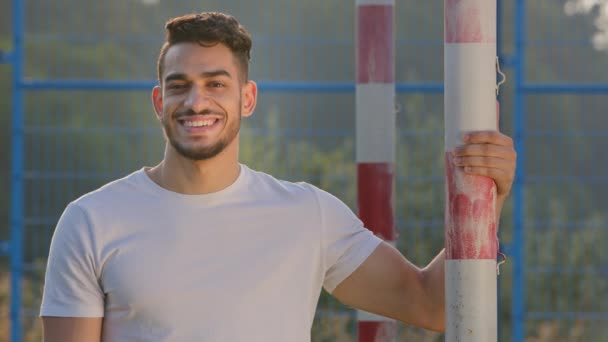 Νεαρός χαμογελαστός αθλητικός προπονητής της Μέσης Ανατολής, τερματοφύλακας ή ποδοσφαιριστής σε καλοκαιρινά αθλητικά που ποζάρουν με φόντο το γκολ στο γήπεδο ποδοσφαίρου. Χιλιετής Άραβας στέκεται στο γήπεδο — Αρχείο Βίντεο