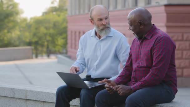 Dwoje starszych ludzi siedzących na zewnątrz, korzystających z laptopa do pracy. Dojrzałe Afroamerykanin męski doradca pomaga stary brodaty kolega lub przyjaciel zainstalować aplikację, budować stronę internetową za pomocą mediów społecznościowych lub napisać e-mail — Wideo stockowe