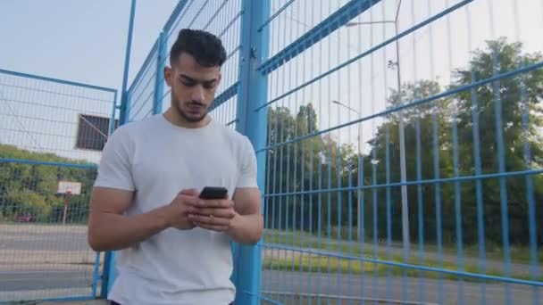 Joven atleta árabe de Oriente Medio en ropa deportiva de verano caminando a lo largo de la valla del estadio sosteniendo el teléfono móvil, utilizando la aplicación en línea en el teléfono inteligente, mensajes de texto, mensaje de escritura de chat, pantalla táctil — Vídeos de Stock