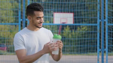 Mutlu gülümseyen Orta Doğu Hindistanlı atlet Latin atlet elinde spor şişeleriyle stadyumda antreman molası sırasında vitamin ve protein karışımı karıştırıyor. Genç adam kardiyo egzersizleri arasında dinlenmeyi seviyor.