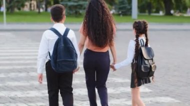 İki çocuk annesi, sırt çantalı kız ve erkek öğrencilerle birlikte şehir caddesinde el ele yürürken arkadan bakan kadın. Anne ve kızı ve oğlu yaya geçidine gidiyorlar.