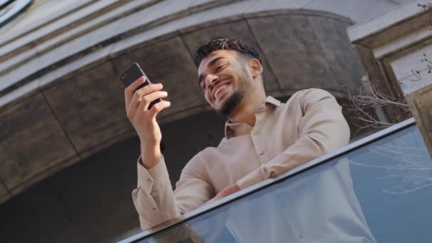 Вид знизу успішний щасливий усміхнений арабський іспано-бородатий багаторічний бізнес-клієнт чоловічої статі, що стоїть на терасі на балконі вдома, тримаючи телефон читає новини отримує повідомлення в додатку Messenger — стокове відео