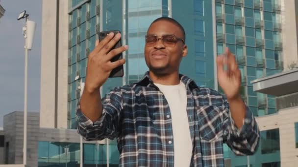 Porträtt av afro amerikan man i solglasögon manliga turist gör videosamtal mobiltelefon app stående i staden visar byggnad bakgrund talar smartphone kamera. Afrikansk kille online konferens utomhus — Stockvideo
