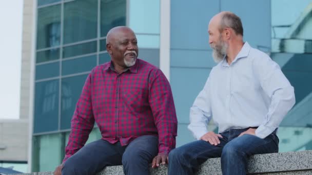 Positif souriant calme hommes âgés afro-américains et européens assis à l'extérieur, avoir une conversation, discuter de questions personnelles ou des problèmes d'affaires. Retraite sans soucis de diversité profiter week-end — Video