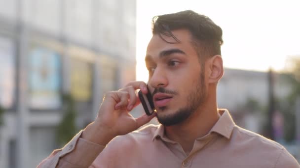 Hombre de negocios hispano barbudo tipo de pie en el fondo de la ciudad de puesta del sol hablando en el teléfono móvil contestando a la llamada de negociación en línea de forma remota. Primer plano conversación cara masculina usando teléfono celular smartphone — Vídeos de Stock