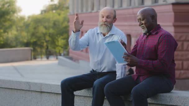 Due persone anziane, maturo adulto afroamericano consulente maschile e vecchio collega barbuto seduto all'aperto. Architetti o artisti esperti ispezionano edifici, analizzano l'architettura, fanno schizzi — Video Stock
