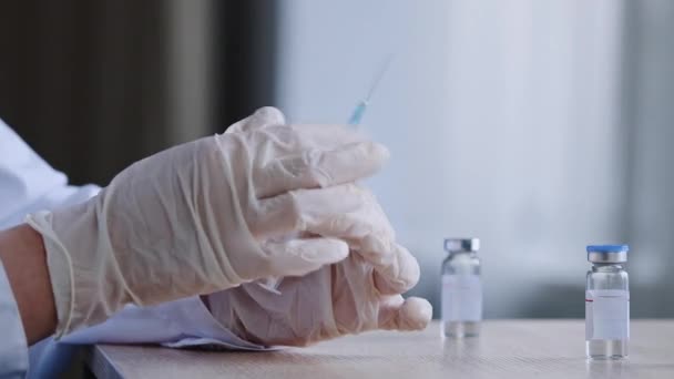 无法辨认的医生护理着身穿白衣和乳胶手套的医护人员 拿着注射器飞溅的药物 准备注射 坐在桌旁 两瓶疫苗和疫苗 — 图库视频影像