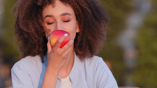 Retrato de niña afroamericana feliz afro mujer de raza mixta étnica dama sentada al aire libre olfateando manzana fragante jugosa roja aplicando fruta a la nariz disfrutando del regreso del aroma después de la enfermedad covid — Vídeos de Stock