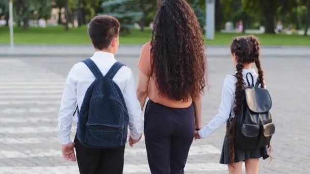 Powrót widok odpowiedzialny matka kobieta trzyma ręce dzieci syn i córka prowadzi uczniów z plecakami do szkoły stanąć na przejściu dla pieszych przejście na drodze rozejrzeć się przestrzegać przepisów ruchu drogowego — Wideo stockowe