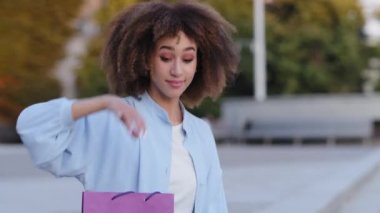 Yakın plan portre alışverişi yapan tüketici kadın tüketici genç Afro-Amerikalı kız model açık havada oturmuş el sallayarak hediye paketini göstermek için arıyor ve gülümsüyor.
