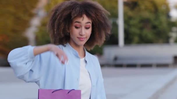 Κοντινό πλάνο πορτρέτο shopper γυναίκα αγοραστής νεαρός αφροαμερικανός κορίτσι θηλυκό μοντέλο κάθεται σε εξωτερικούς χώρους πόλη κουνώντας το χέρι καλώντας να έρθει δείχνει φωτεινό τσάντα δώρο με την παρούσα αγορά αγορών χαμογελώντας — Αρχείο Βίντεο