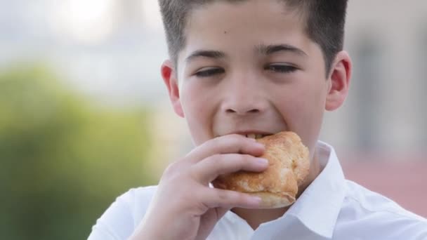 Portré gyermek arc spanyol kisfiú iskolás fiú kis tanuló áll a szabadban napsugarak eszik harapás finom puha zsemle élelmiszer sütés élvezi eszik édesség éhség iskola után leckék nézi a kamera — Stock videók