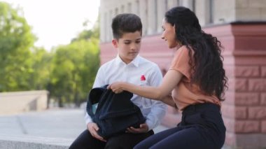 Değer veren genç anne anne anne okuldan sonra oğluyla dışarıda oturur, anne çocuk çengelli yılan tişörtünü düzeltir, sırt çantasına su koyar, çocuğun kafasını okşayıp sohbet etmesine yardım eder.