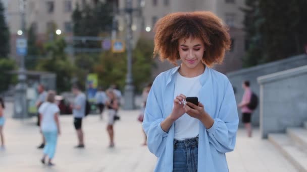 Junge lockige Frau ethnische Mädchen afrikanisch-amerikanische Studentin, die in der Stadt im Freien steht und auf den Telefonbildschirm schaut, benutzt Handy für Chats im Netz mit winkendem Kopf — Stockvideo