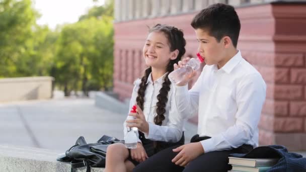 Dos niños escolares compañeros de clase hispanos hermano y hermana niña y niño niños con camisas blancas sentados al aire libre después de la escuela hablando riendo bebiendo agua fría y limpia de una botella de plástico — Vídeos de Stock