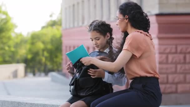 Familia hispana niña linda niña niña colegiala con mamá sentado al aire libre conversando conversación comunicación charla preparándose para la escuela poner cuaderno libro en la mochila — Vídeos de Stock
