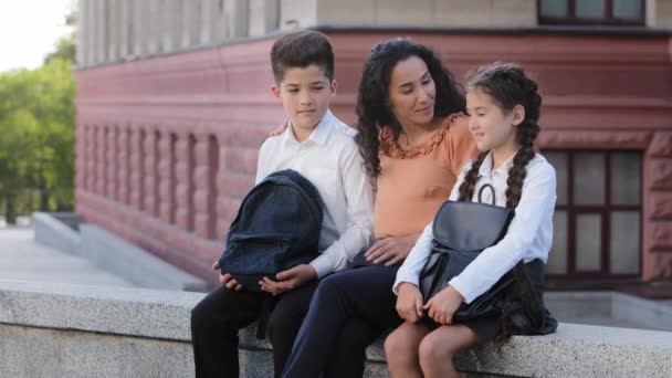 Mujer joven madre cariñosa madre maestra se sienta en el patio de la escuela en la calle al aire libre con los niños amados hija e hijo escolares hablan, familia hispana dos estudiantes y mamá hablando después de la escuela — Vídeos de Stock