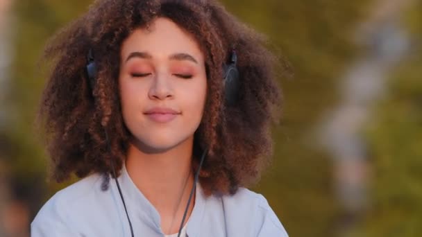 Портрет красивої щасливої афроамериканської дівчини афро-жінки етнічної змішаної раси леді, яка сидить на відкритому повітрі в місті, носить навушники на голові, слухаючи музику, насолоджуючись пісенною мелодією слухати аудіо — стокове відео