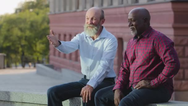 Divertidos alegres y despreocupados ancianos de edad de jubilación sentados al aire libre en la ciudad disfrutando de fin de semana, coqueteando juguetón. Dos diversos amigos caucásicos europeos y afroamericanos divirtiéndose juntos — Vídeo de stock