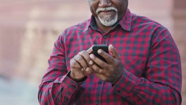 Starszy Murzyn w średnim wieku trzymający nowoczesny smartfon za pomocą aplikacji mobilnej, starsza osoba męskie ręce piszące sms-y online przez telefon komórkowy, starsi ludzie i koncepcja technologii, widok z bliska — Wideo stockowe
