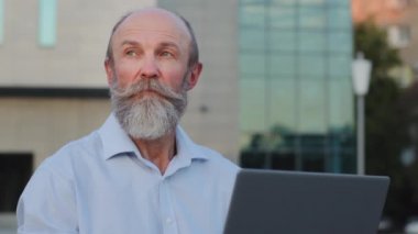 Yakın plan, gri sakallı yaşlı adam dışarıda dizüstü bilgisayar kullanıyor. Ciddi emekliler uzaktan kumandalı çalışır. Yaşlı insan emekli olduğunda modern teknoloji ve bilgisayar uygulaması kullanır.