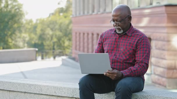 Serious focused senior black person using laptop, African american mature male communicating on social networks online, elderly man writing emails, distantly working on computer in public place — Stock Video