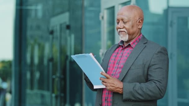 Ancien sérieux concentré homme d'affaires, architecte ou artiste debout à l'extérieur tenant du papier presse-papiers inspecte les bâtiments, afro-américain spécialiste masculin analyse l'architecture, fait des croquis — Video