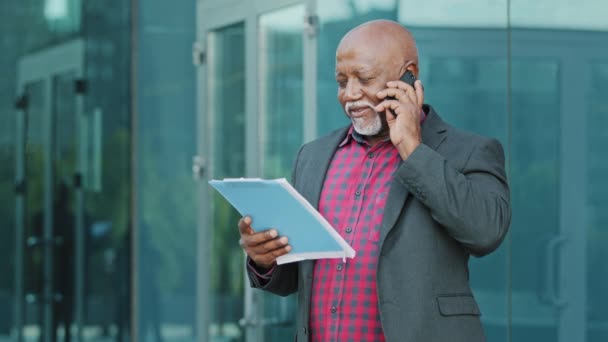 Επιχειρηματίας ή αρχιτέκτονας κατέχει smartphone, συζητά λεπτομέρειες του έργου με τον πελάτη ή το αφεντικό στο τηλέφωνο, έχει επιχειρηματική συνομιλία. Ώριμος Αφρικανός ειδικός στέκεται σε εξωτερικούς χώρους με πρόχειρο χαρτί — Αρχείο Βίντεο