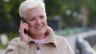Mutlu orta yaşlı, Kafkasyalı yaşlı kadın büyükannem çocuklarla uzaktan akıllı telefondan konuşarak şehir parkında hafta sonunun tadını çıkarıyor. Yakın plan kadın yüzü yaşlı bayan cep telefonundan haberleri paylaşıyor