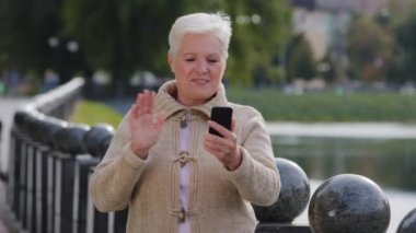 Aşırı neşeli Kafkasyalı yaşlı kadın el sallıyor akıllı telefon kamerasına bakıyor modern cep telefonuyla konuşuyor, mutlu orta yaşlı bayan konuşuyor keyifli bir iletişimi var
