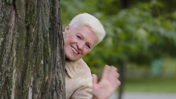 Neşeli olgun orta yaşlı kadın ağacın arkasına saklanıyor, oyun oynuyor, eğleniyor, el sallıyor, gülümsüyor yaşlı bayan, mutlu, emekli, güzel büyükanne haftasonu dışarıda parkta eğleniyor. — Stok video
