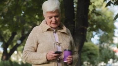 Gülümseyen, yaşlı, gri saçlı, emekli bir kadın. Elinde plastik bir fincan kahve ve telefonla parkta yürüyor. Mutlu yetişkin kadın emeklilik yaşı akıllı telefon ekranına bakıyor açık havada postaları kontrol ediyor.