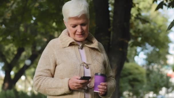 Uśmiechnięta, atrakcyjna emerytka z siwymi włosami, chodząca po parku z plastikową filiżanką kawy i telefonu. szczęśliwy dorosły Dojrzałe lady wiek emerytalny patrząc na ekran smartfona sprawdzanie poczty outdoor — Wideo stockowe