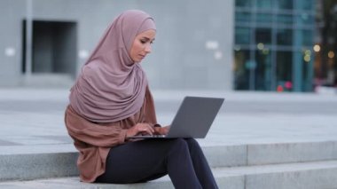 Ciddi odaklı müslüman genç iş kadını serbest çalışan kadın tesettürlü kız tesettüre girmiş kadın kullanıcı dışarıda dizüstü bilgisayarda oturup uzaktan öğrenme için bilgisayar uygulaması kullanıyor.