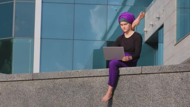 Мусульманская индийская женщина в хиджабе девушка йоги гимнастка сидит в городе с ноутбуком работает онлайн держа ногу за головой растягивая неудобную позу акробатическая асана йога гибкость — стоковое видео