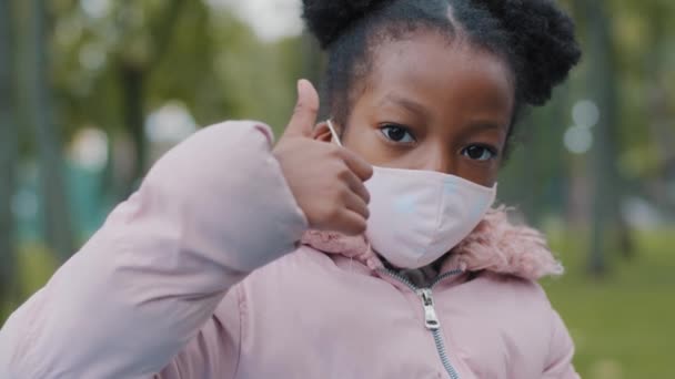 Портрет в городе маленькая серьезная африканская девочка афро ребенок стоит на улице, глядя в камеру показывает большие пальцы вверх рекомендует носить медицинскую маску карантина коронавируса ковид пандемии — стоковое видео