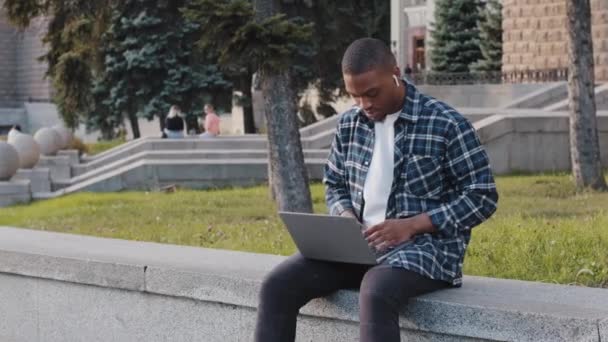 Успішний зайнятий африканський молодий чоловік афро хлопець студент чоловік фрілансер дизайнер в бездротових навушниках, що сидять на терасі в місті в парку, друкуючи ноутбук балачки, що працює дистанційно онлайн — стокове відео